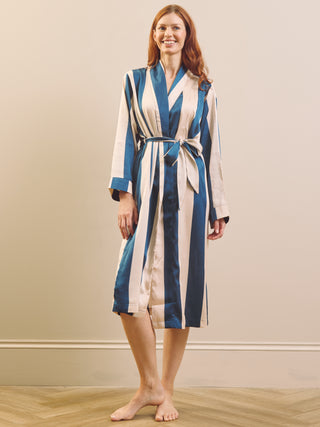 A woman wearing a long-sleeved striped bathrobe with blue and cream vertical stripes, standing barefoot on a light wood floor against a beige wall.