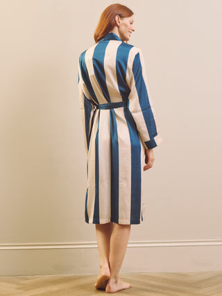 A woman stands barefoot indoors wearing a long-sleeve striped robe with vertical blue and cream stripes and a matching tie belt, shown from the back.