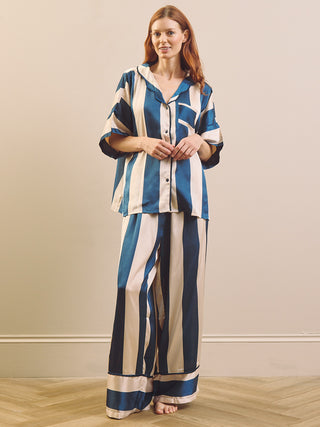 A woman stands barefoot in a room wearing a matching striped pyjama set with a button-front long-sleeve top and wide-leg pants. The fabric has vertical stripes in blue, navy, and cream.