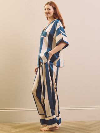 A woman modelling a matching two-piece pyjama set with vertical stripes in navy blue and cream, standing barefoot against a neutral beige wall.
