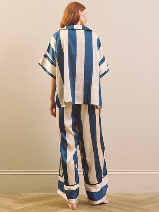 A woman stands barefoot indoors, viewed from the back, wearing a loose matching pyjama set with vertical blue and off-white stripes. The set includes a short-sleeve collared top and wide-leg pants.