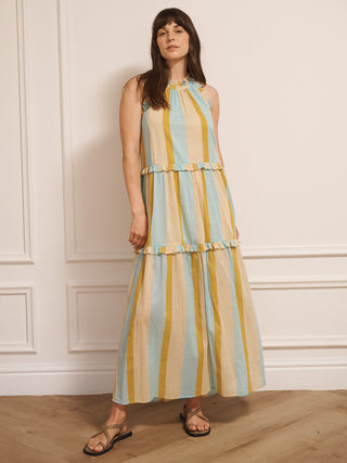 Model wearing an aqua and gold striped cotton tiered halter neck maxi dress with frill detailing, standing indoors on a wooden floor.