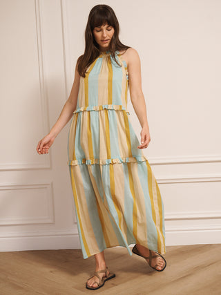 Model wearing an aqua and gold striped cotton tiered halter neck maxi dress with frill detailing, standing indoors on a wooden floor.