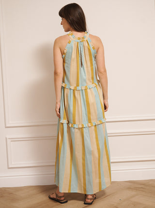 Model wearing an aqua and gold striped cotton tiered halter neck maxi dress with frill detailing, standing indoors on a wooden floor. Back detail