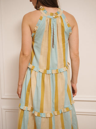 Model wearing an aqua and gold striped cotton tiered halter neck maxi dress with frill detailing, standing indoors on a wooden floor.