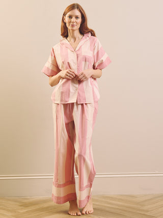 A woman stands barefoot indoors wearing a pink striped pyjama set consisting of a short-sleeve button-front top and matching loose pants.