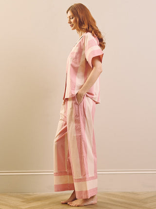 A woman stands in profile wearing a light pink triped pyjama set consisting of a short-sleeve top and matching wide-leg pants, set against a neutral beige wall and light wood herringbone floor.