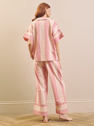 A woman stands barefoot in a room with a beige wall and light wood herringbone floor, viewed from behind. She is wearing a pink striped short-sleeve pyjama top and matching wide-leg pyjama pants.
