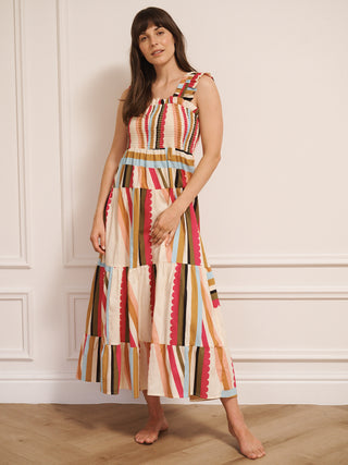 Model wearing a multicolour striped cotton tiered dress with shirred bodice and wide ruffled straps, standing barefoot indoors on a wooden floor.