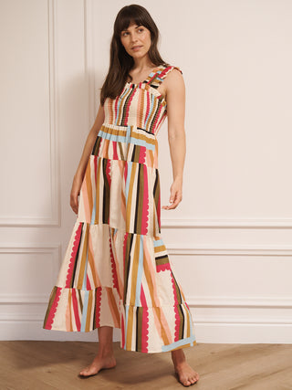 Model wearing a multicolour striped cotton tiered dress with shirred bodice and wide ruffled straps, standing barefoot indoors on a wooden floor.