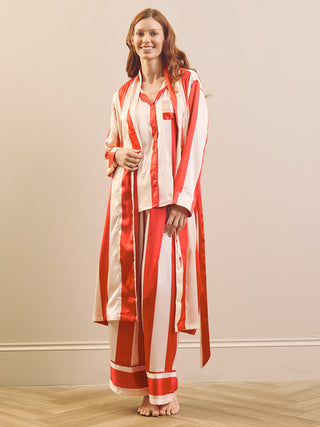 A woman modelling a matching red and white striped pyjama set with a long dressing gown robe, standing barefoot on a light wood floor against a beige wall.