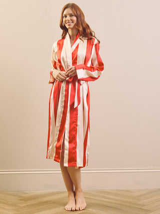 A woman wearing a red and white vertically striped silk-like robe with a matching belt, standing barefoot on a light wood floor against a plain beige wall.