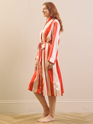 A woman wearing a satin-like robe with vertical red and cream stripes, standing barefoot in a room with a beige wall and light wood floor.
