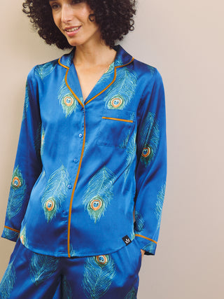 Woman wearing a blue satin pajama set with peacock feather design against a beige background
