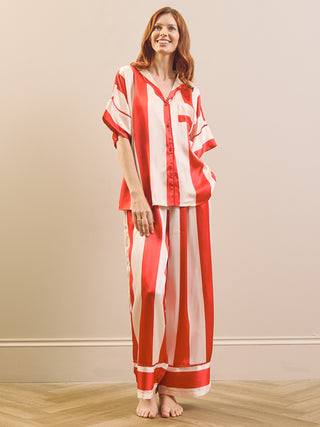 A woman modelling a matching two-piece pyjama set with vertical red and white stripes, featuring a short-sleeve button-front top and wide-leg pants, standing against a neutral beige wall.