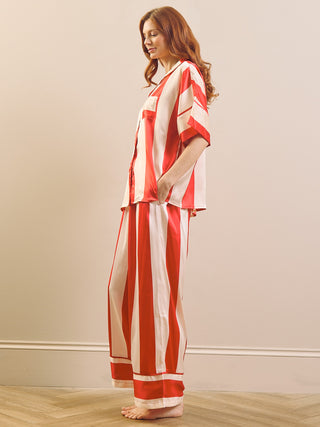 A woman modelling a matching two-piece pyjama set with bold red and white vertical stripes, standing in profile against a neutral beige wall and light wood floor.