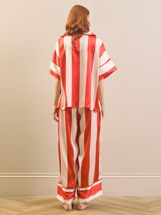 A woman stands with her back to the camera wearing a matching two-piece pyjama set with bold red and white vertical stripes. The set includes a short-sleeve top and wide-leg pants, photographed against a neutral beige wall and light wood floor.