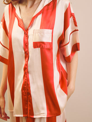 A close-up of a person wearing a short-sleeve satin button-front pyjama shirt with bold vertical red and off-white stripes and a chest pocket, against a plain light beige background.