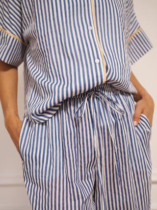 Model wearing women’s navy ticking stripe cotton gauze pyjamas with short-sleeved button-up top, drawstring waist trousers and contrast trim, photographed in a neutral studio setting. Tie waist detail
