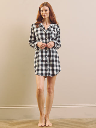 A woman stands barefoot against a beige wall wearing a black and white gingham plaid long-sleeve button-up nightshirt dress.