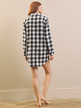 A woman with red hair is shown from behind wearing a black and white gingham check plaid long-sleeve nightshirt dress, standing barefoot on a light wood floor against a beige wall.