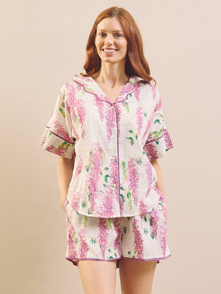 A woman stands barefoot in a bedroom setting wearing a matching short-sleeve button-front pyjama top and shorts set with a pink and green floral print on a white background.