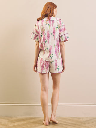 A woman stands barefoot indoors facing away from the camera, wearing a matching short-sleeve pyjama top and shorts set with a pink floral print on a light cream background.