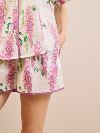 A close-up of a person wearing a matching pyjama set with a wisteria floral print. The set features a short-sleeve button-up top and shorts in an off-white fabric with pink, purple, and green wisteria flowers and leaves, finished with a purple piping trim.
