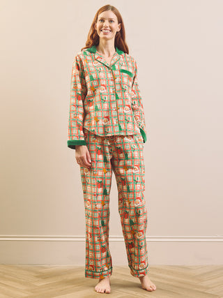A woman stands barefoot in a bedroom setting wearing a matching two-piece pyjama set. The pajamas feature a multicolor plaid pattern with green accents and a subtle sheen, set against a neutral beige wall and light wood floor.