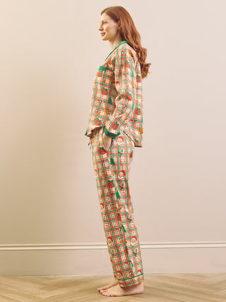 A woman stands in profile wearing a long-sleeve pyjama set with a colorful geometric pattern in orange, green, and white, set against a neutral beige wall and light wood floor.