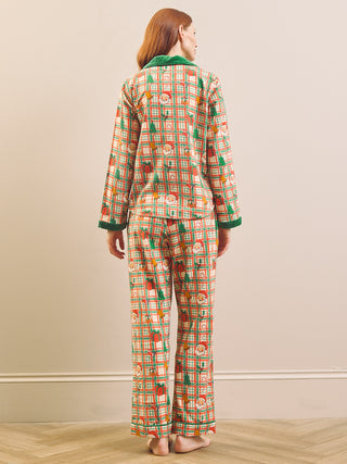 A woman stands barefoot indoors wearing a matching long-sleeve pyjama set shown from the back. The pajamas have a multicolor holiday-themed print with green trim on the collar and cuffs, set against a light beige wall and pale wood floor.