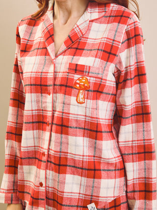 A close-up of a person wearing a red, white, and black tartan plaid button-up shirt with a small embroidered orange-and-white mushroom patch on the chest pocket.