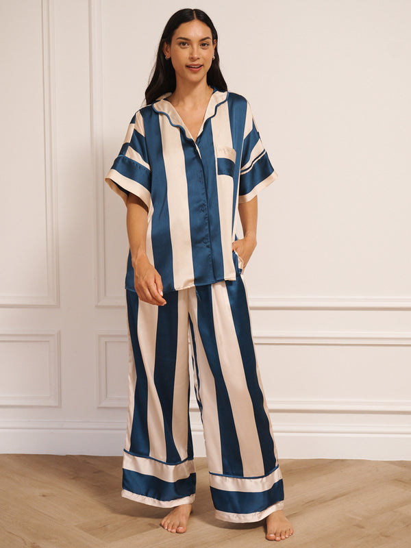 Model wearing women’s navy and cream striped satin oversized pyjamas with short-sleeved button-up top and wide-leg trousers, photographed barefoot in a neutral studio setting.