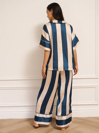 Model wearing women’s navy and cream striped satin oversized pyjamas with short-sleeved button-up top and wide-leg trousers, photographed barefoot in a neutral studio setting. back shot