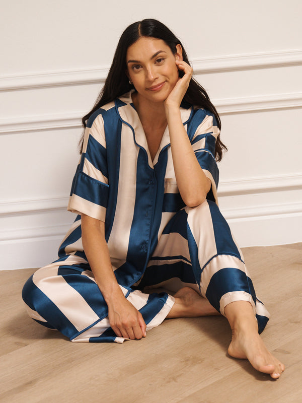 Model wearing women’s navy and cream striped satin oversized pyjamas with short-sleeved button-up top and wide-leg trousers, photographed barefoot in a neutral studio setting sat on the floor