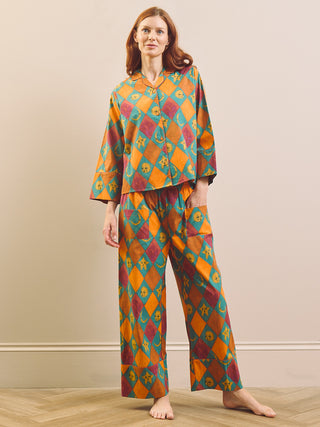A woman stands barefoot indoors wearing a matching two-piece pyjama set with a bold geometric harlequin pattern in orange, teal, burgundy, and yellow. The set includes a long-sleeve button-front top and wide-leg pants.