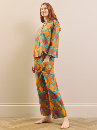 A woman stands barefoot in a room wearing a matching two-piece pyjama set with a colorful geometric ikat pattern in orange, teal, and burgundy.