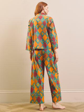 A woman stands barefoot indoors wearing a matching two-piece pyjama set with a colorful harlequin diamond pattern in orange, teal, and burgundy. The set includes a long-sleeve top and wide-leg pants, shown from the back against a plain beige wall.