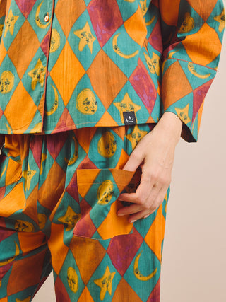 A close-up of a person wearing a colorful patterned pyjama set with a diamond print in orange, teal, and magenta, featuring yellow celestial motifs like stars and crescent moons. The person is shown from the chest down, with one hand pulling the pocket of the pants.