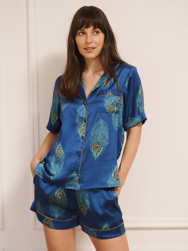 Model wearing a blue satin short pyjama set with peacock feather pattern against a white background
