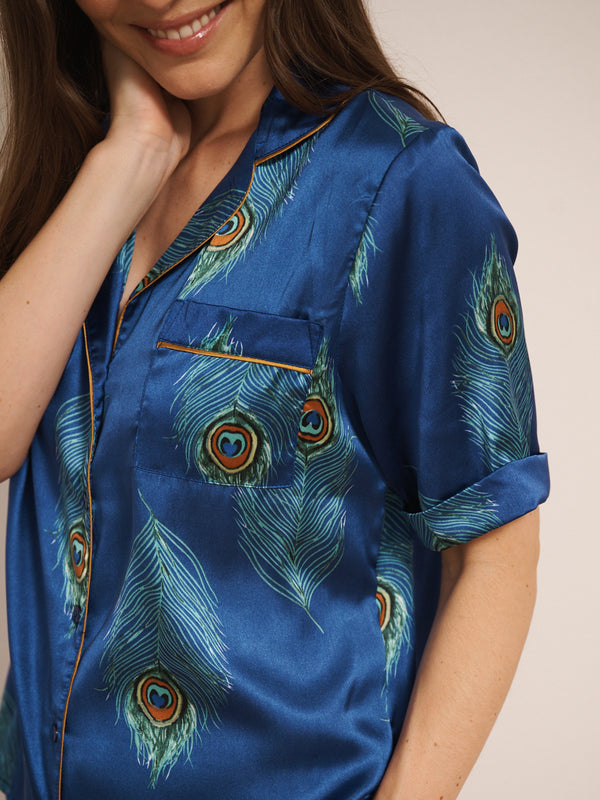 Model wearing a blue satin short pyjama set with peacock feather pattern against a white background