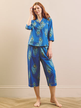 A woman modelling a two-piece pyjama set with a vibrant blue satin-like fabric and a peacock feather print. The set includes a short-sleeve button-up top and matching cropped pants, photographed against a neutral beige wall and light wood floor.