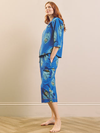 A woman stands barefoot in profile wearing a matching pyjama set with a bright blue base and a peacock feather print. The set includes a short-sleeve top and cropped pants, photographed against a plain beige wall and light wood floor.