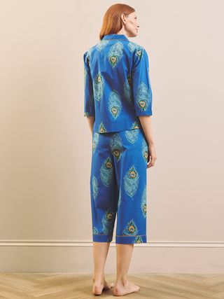 A woman stands barefoot against a beige wall wearing a matching blue pyjama set with a peacock feather print. The set includes a short-sleeve button-up top and cropped pants.