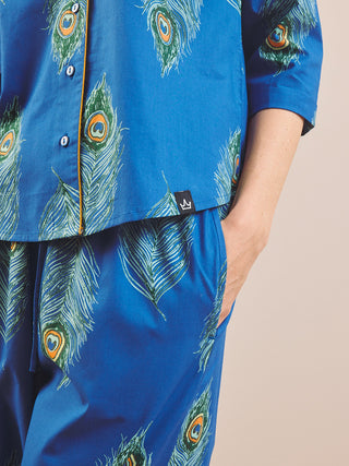 A close-up of a person wearing a blue pyjama set with a peacock feather print. The set includes a short-sleeve button-front top and matching pants, with a small black brand tag visible on the hem.