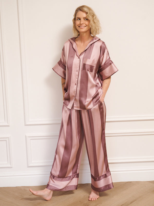 Model wearing women’s mauve and blush striped satin oversized pyjamas with short-sleeved button-up top and wide-leg trousers, photographed barefoot in a neutral studio setting.