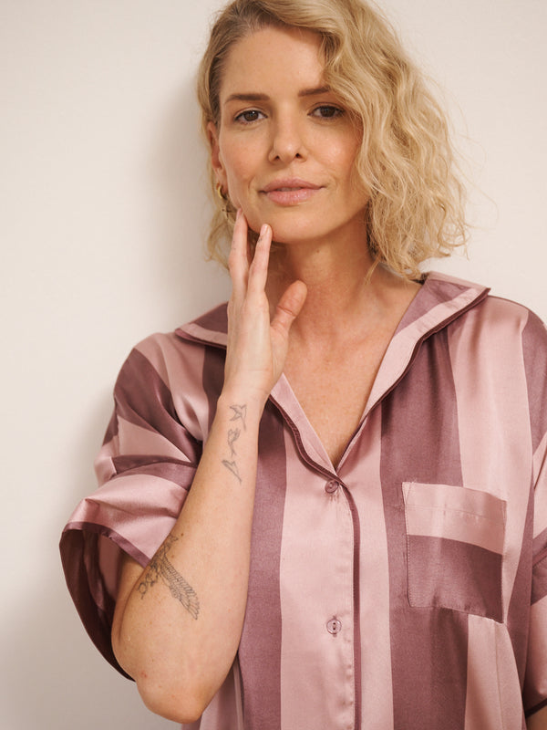 Model wearing women’s mauve and blush striped satin oversized pyjamas with short-sleeved button-up top and wide-leg trousers, photographed barefoot in a neutral studio setting. close up. 