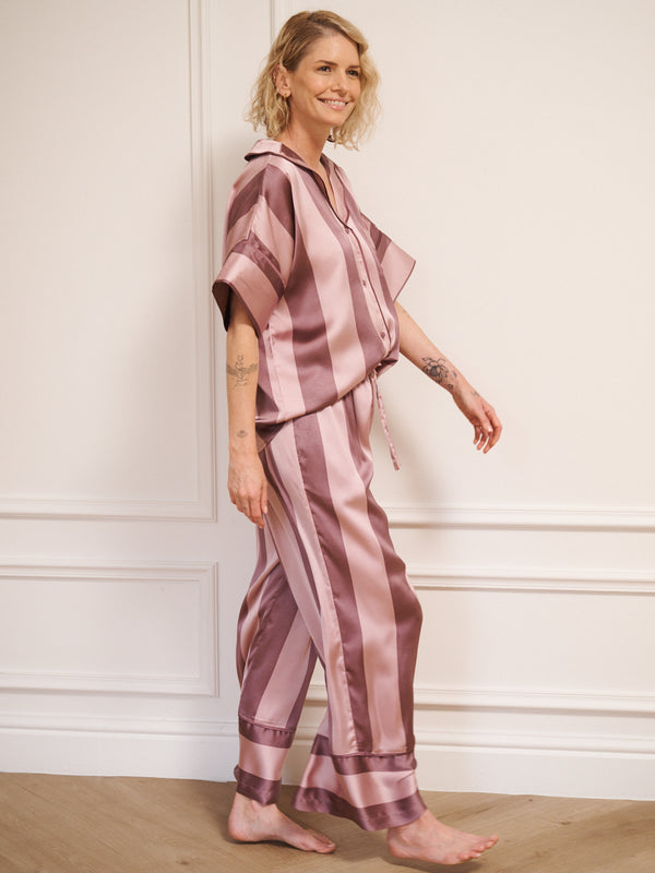 Model wearing women’s mauve and blush striped satin oversized pyjamas with short-sleeved button-up top and wide-leg trousers, photographed barefoot in a neutral studio setting. Side Detail. 