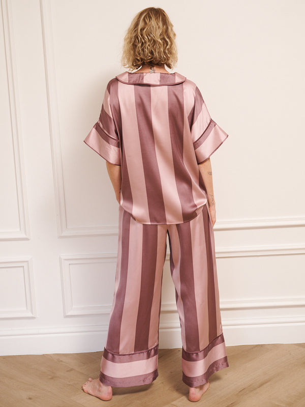 Model wearing women’s mauve and blush striped satin oversized pyjamas with short-sleeved button-up top and wide-leg trousers, photographed barefoot in a neutral studio setting. Back detail