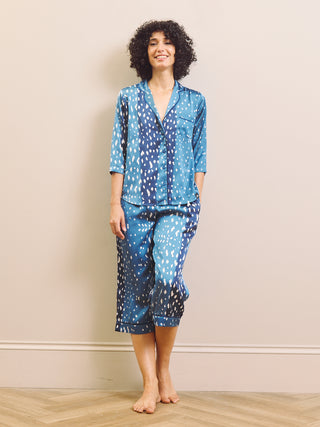 A woman modeling a two-piece capri pyjama set with a blue and white abstract pattern, standing barefoot against a beige wall and light wood floor.